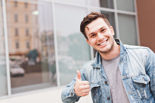 Young successful smiling man businessman in casual jeans jacket showing thumbs up gesture outdoors on street in summer in front of mirror glass office building or business center