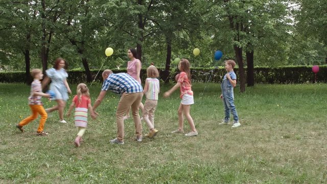 Little boys and girls playing dodge ball with their parents on green lawn decorated for birthday party