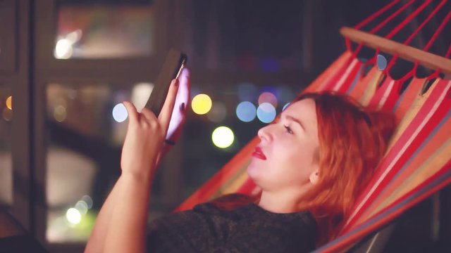 Beautiful woman with red hair using smart phone mobile in the city at night liying in hammock. 1920x1080