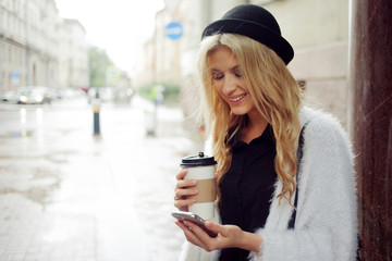 Cheerful woman in the street drinking morning coffee and use her smartphone.