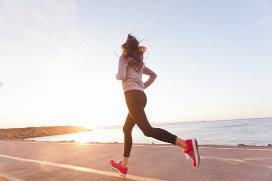 Young woman athlete running at sunset on seashore sidewalk. Girl runner training on a beach