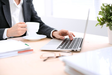 Close up of business woman  hands  with a cup of coffee is sitting at the table and typing on a laptop computer in the white colored office .