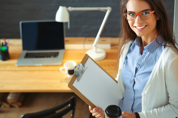Young woman standing near desk with laptop holding folder and cup of coffee