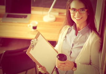 Young woman standing near desk with laptop holding folder and cup of coffee