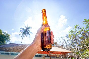 Vacation concept. Male hand holding bottle of beer against sunny sky.