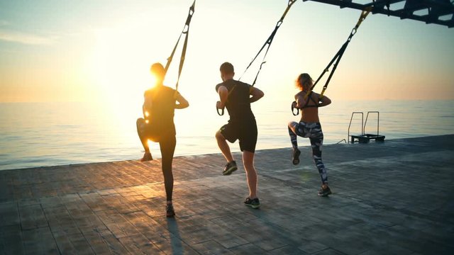 Sportive people training squats with trx at seashore against sun. Slow motion.