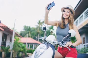Obraz premium Summer lifestyle and technology. Outdoor portrait of pretty young woman in hat sitting on scooter taking selfie on her smart phone.