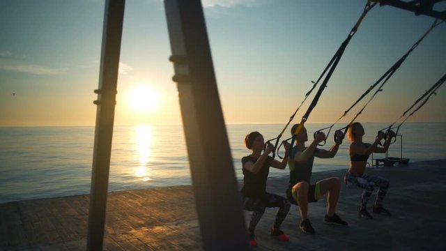 Silhouettes of sportive people training squats with trx at sunrise. Slow motion.