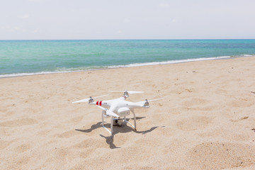 Fototapeta na wymiar White Drone with four propellers standing on a sandy, bright beach in the sunshine. The blue sea in in the background