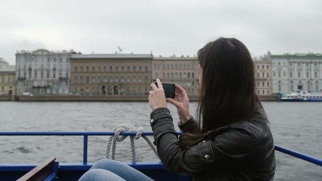 Girl sitting in a river bus on the go taking photos of architecture on a river quay. A seagull is flying around, slow mo