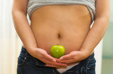 Closeup womans stomach with unzipped jeans and shirt lifted up, holding apple between hands, underwear visible, weightloss concept