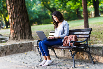 Young female student sitting on a bench in park, she is working on laptop.