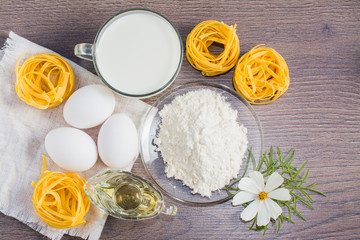 Лапша, куриные яйца, пшеничная мука, подсолнечное масло, молоко и садовый цветок на деревянном фоне