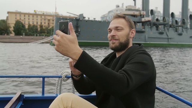 Happy handsome man sightseeing in St Petersburg. Taking selfie in front of Cruiser Aurora, uses his smartphone, slow mo