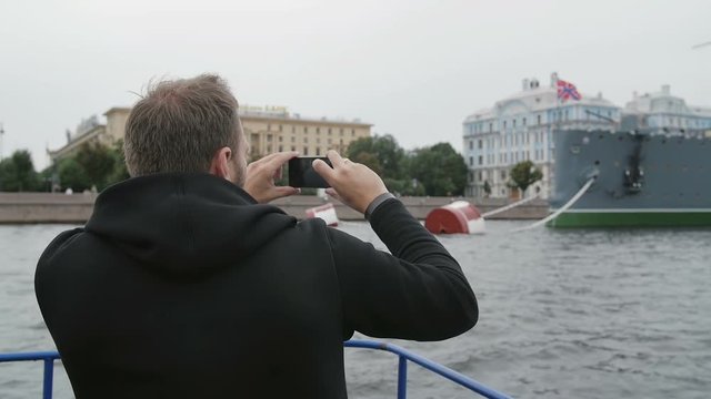 Sightseeing in St Petersburg. Man uses smartphone, takes photos of the Cruiser Aurora, passing by it on a boat, slow mo