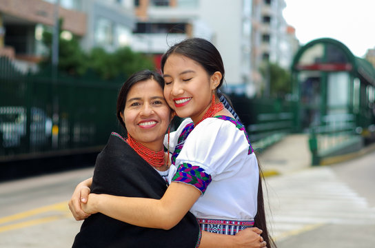Beautiful hispanic mother and daughter wearing traditional andean clothing, waiting for bus at public station while embracing together, smiling happily, outdoors environment