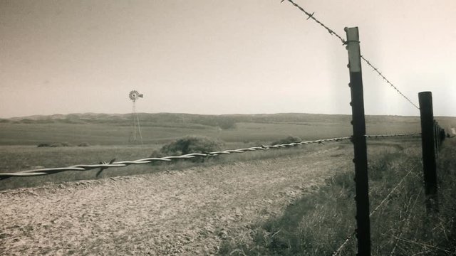 A vintage styled perspective shot looking through a barded wire fence at a wind mill on a farm.