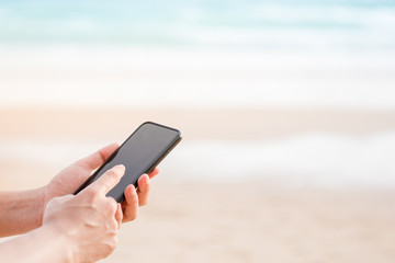 left hand holding mobilephone, index finger of right hand pointing at screen, blurred beach on background