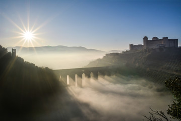 Spoleto (Italy) - A misty fall day in the charming medieval village in Umbria region. The soft focus depends on dense fog, which, however, creates an evocative atmosphere with Sun rays  - obrazy, fototapety, plakaty