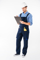 Pensive young builder in helmet thinking and writing on clipboard