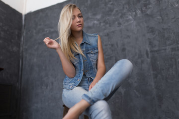 Portrait of the blonde in jeans and denim sleeveless jacket with her hair and natural makeup sitting on a wooden chair