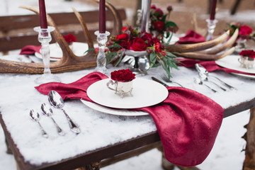 Wedding table setting in marsala colors with plates, cutlery, red floral compositions, candles, velvet napkins, deer horns on table covered with snow