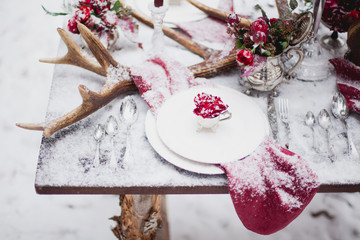 Wedding table setting in marsala colors with plates, cutlery, red floral compositions, candles, velvet napkins, deer horns on table covered with snow