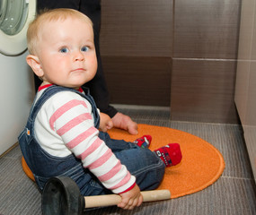 Funny 1 year old boy is holding a plunger sitting on the floor in a bathroom near washing machine - 122125682