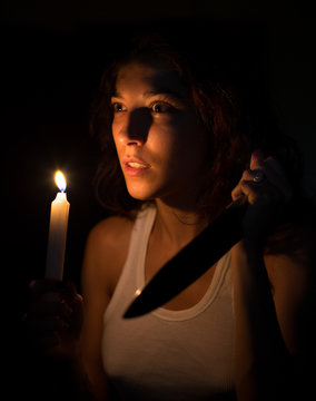 Mujer joven mirando asustada en un apagón iluminada por una vela con un cuchillo en la otra mano