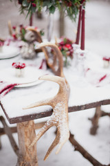 Wedding table setting in marsala colors with plates, cutlery, red floral compositions, candles, velvet napkins, deer horns on table covered with snow