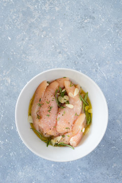 Raw chicken tenders marinated with garlic, olive oil, lemon juice, rosemary, thyme, salt and pepper in white bowl