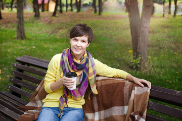Outdoor fashion details. Lifestyle image of stylish young attractive woman in cozy knitted sweater , holding cup of hot beverage , sitting on wooden city bench.
