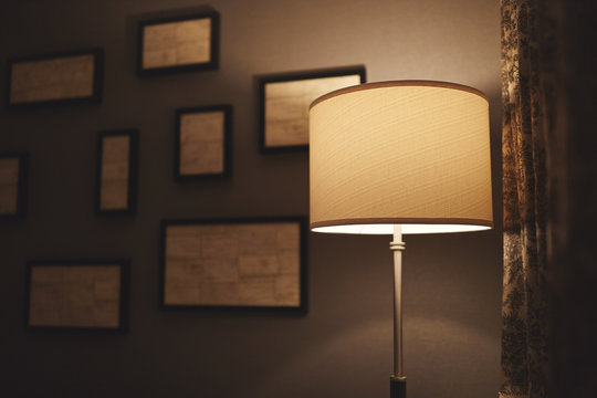 A Lamp In A Dark Corner Of A Hotel Room With Abstract Frames Out Of Focus On The Wall Behind It And A Curtain To The Right.