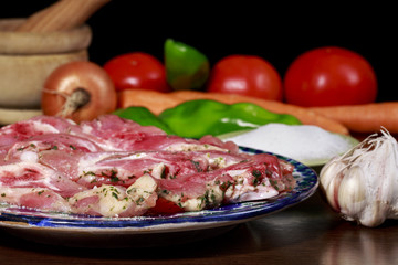 Dish with raw turkey meat marinated with garlic and parsley, beside garlic bulb, small dish with salt, green peppers and other vegetables. On black background
