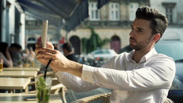 Handsome, stylish man doing selfies on tablet while sitting in the outdoor cafe
