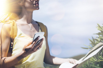 Hipster young girl with bright backpack looking at phone and map. Said view tourist traveler on background mountain, blue sea, sun flare. Mockup for text message. Female hands using smartphone