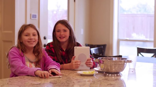 Two friends sitting at a kitchen island using their tablets and smiling at the camera
