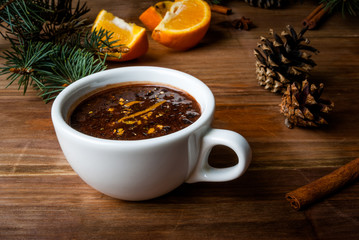 Hot chocolate with spices, orange and orange zest. Against the background of Christmas tree branches, cones and orange. Wooden table