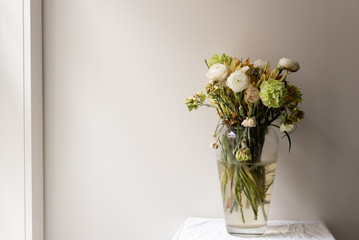 Wilting green and white ranunculus and carnations in large glass vase on small white table next to window