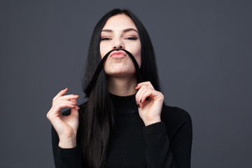 Fototapeta premium Young brunette woman is playing with long hair and making mustache of it, gray background