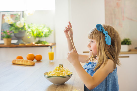 SIDE VIEW: Little girl in a blue dress funny eats a spaghetti from a dish