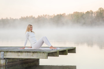 Woman relaxing and practicing yoga in the mist on the lake footbridge early morning. White clothes.