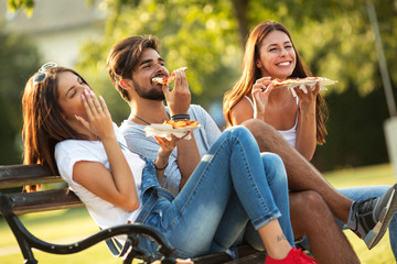 Group of young friends sitting on park bench and eating pizza.Fast food.