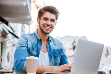 Smiling casual man using laptop while sitting at cafe outdoors