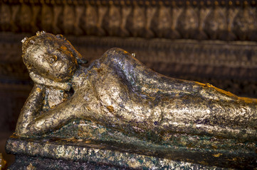 Small Buddhist statue covered in gold leaf reclines in front of a golden textured background at a Buddhist temple in Bangkok, Thailand