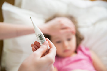 Close-up thermometer.  Mother measuring temperature of her ill kid. Sick child with high fever laying in bed and mother holding thermometer. Hand on forehead.  - obrazy, fototapety, plakaty