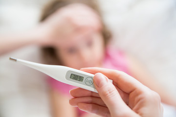 Close-up thermometer.  Mother measuring temperature of her ill kid. Sick child with high fever laying in bed and mother holding thermometer. Hand on forehead.  - obrazy, fototapety, plakaty