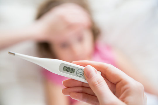 Close-up thermometer.  Mother measuring temperature of her ill kid. Sick child with high fever laying in bed and mother holding thermometer. Hand on forehead. 