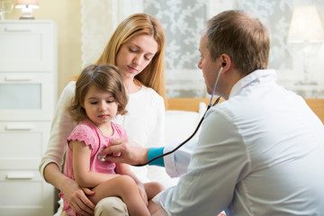 Male Pediatrician examining cute little girl with stethoscope. Kid looks sick and sad. Mother holding her kid. Doctor visit his patient at home.