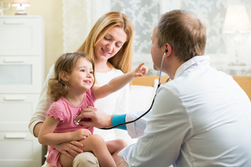 Male Pediatrician examining cute little girl with stethoscope. Kid happily smiling and pointing on...